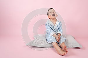 Cranky child boy refuses to go to bed. Kid boy in bathrobe sits on pillow on pink background