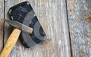 Crashed black smartphone is lying on the old wooden background. Broken lcd touch screen with a hammer. Information technology