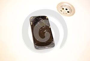 Crashed black smartphone in lying in the white wash basin sink. Broken lcd touch screen in the bathroom. Information technology