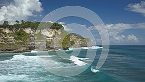 Crashing ocean waves carry lonesome boat towards towering cliff on a calm day.