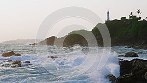 Crashing waves hit rocky shore near white lighthouse. Palm trees sway as surf surges around stones. Misty sea spray