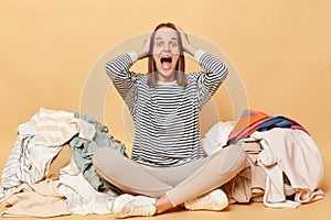 Crazy amazed Caucasian woman posing near heap of multicolored unsorted clothes isolated over beige background needs sorting attire