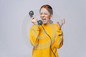 Crazy angry young woman in stylish yellow sweater talking on retro phone and screaming in handset.