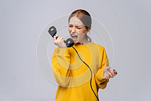 Crazy angry young woman in stylish yellow sweater talking on retro phone and screaming in handset.