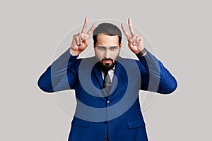 Crazy bearded man showing bull horn gesture with fingers over head, looking hostile and threatening.