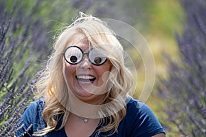 Crazy Blonde woman wearing googly eyes novelty sunglasses while sitting in a field of lavender, looking surprised with her mouth