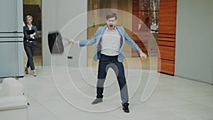 Crazy businessman dancing with briefcase in modern lobby while his colleagues walking and watching him surprised