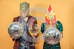 Crazy couple having fun celebrating new year eve party holding disco balls and glass of wine - Focus on t-rex and chicken mask
