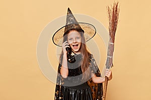 Crazy excited little girl with long hair black cloak clothing with broom isolated over beige background talking on mobile phone