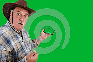 A crazy farmer looks directly at the camera while holding a smartphone with a green screen in his hand.