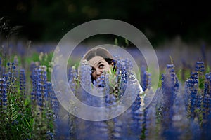 Crazy funny girl with beautiful smilie hiding in green field with many flowers, lupins in summer. HIDING CONCEPT