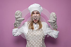 Crazy girl cook in uniform shouts and raises hands and shows baking gloves on a colored background, woman housewife in kitchen