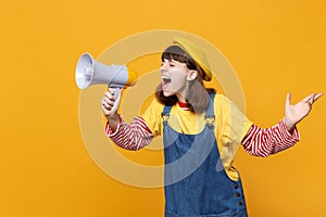 Crazy girl teenager in french beret and denim sundress looking aside, scream in megaphone isolated on yellow wall