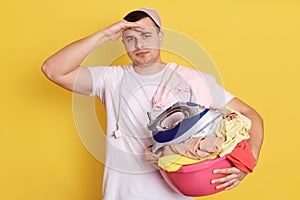 Crazy househusband holding iron near ear like talking via smart phone, holding basin with clothing for laundry, posing isolated
