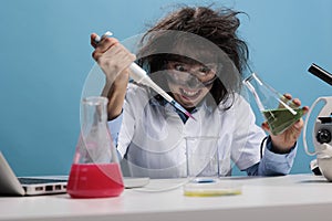 Crazy mad scientist mixing liquid substances while grinning dreadful and sitting at desk on blue background.