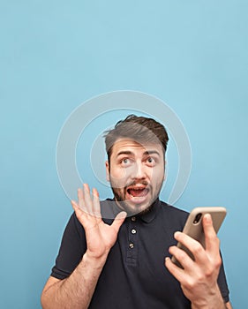 A crazy man with a beard standing on a blue background with a smartphone in his hands, with a shocking face and an open mouth