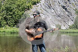 crazy man musician, guitarist on vacation. 23 year guy with beard in black shirt, cap, sunglasses holds steaming guitar