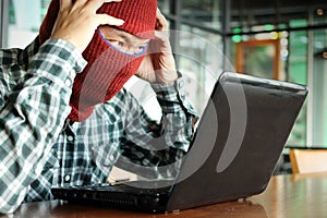 Crazy masked hacker wearing a balaclava touching on head between stealing data from laptop. Internet crime concept.