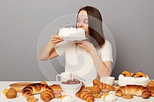 Crazy woman with brown hair wearing white T-shirt sitting at table  over gray background, bites hungrily delicious sweet