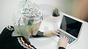 Crazy woman typing on laptop with foil hat to shields her from 5G waves, internet, electromagnetic fields, mind control