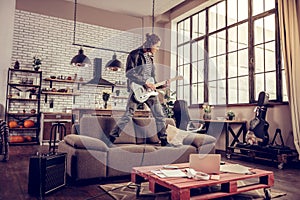 Crazy young rock musician standing on sofa and playing music