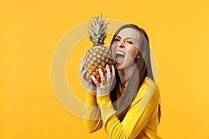 Crazy young woman in casual clothes holding, biting fresh ripe pineapple fruit isolated on yellow orange wall background