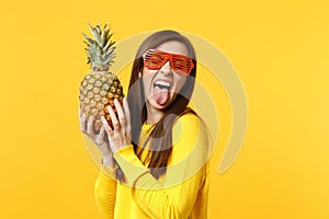 Crazy young woman in funny glasses showing tongue, holding in hands fresh ripe pineapple fruit isolated on yellow orange