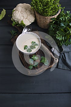 Cream of cauliflower soup with cream and greens, in a white cup on a wooden board and black background. Vintage photo. Fresh