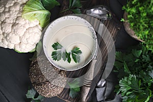 Cream of cauliflower soup with cream and greens, in a white cup on a wooden board and black background. Vintage photo. Fresh