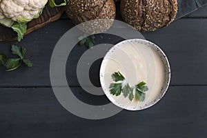 Cream of cauliflower soup with cream and greens, in a white cup on a wooden board and black background. Vintage photo. Fresh