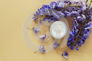 Cream jar and purple wisteria flowers on beige background. Natural organic cosmetics concept. Top view, flat lay, copy