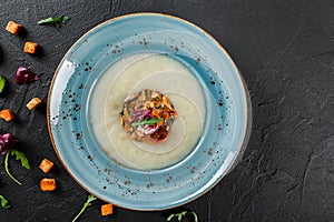 Cream soup of celery with tartare from baked vegetables decorated from croutons and arugula in plate on dark stone background