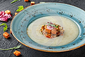 Cream soup of celery with tartare from baked vegetables decorated from croutons and arugula in plate on dark stone background