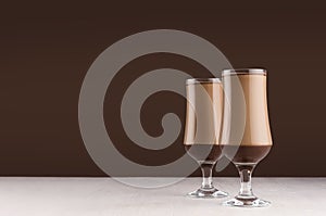 Creamy coffee cocktail in two elegant glass with chocolate on white table and dark brown wall.