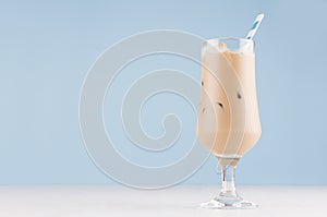 Creamy cold fresh beverage in elegant transparent glass with ice cubes and striped straw in modern light blue interior.