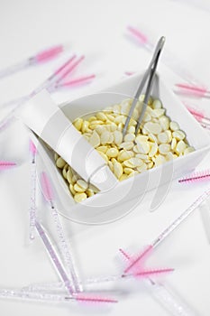 Creamy colored depilatory wax beans lying in bowl with tweezers and tube surrounded by eyelash brushes on white table.