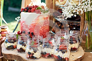 Creamy dessert with fresh berries in a glass jar. Desserts stand on a wooden block, with a birthday cake in the background.