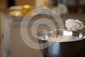 Creamy egg whites on mixer whisks over bowl with kitchen in background