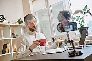 Creating content. Cheerful male blogger making a new video for his vlog and drinking a tea while sitting indoors.