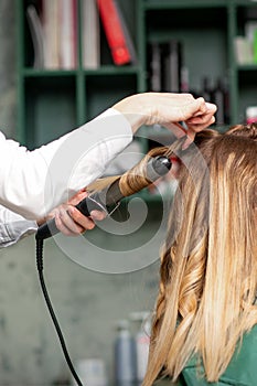 Creating curls with curling irons. Hairdresser makes a hairstyle for a young woman with long red hair in a beauty salon.
