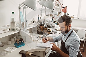 Creating sketches. Portrait of young bearded jeweler drawing a sketch of a new project