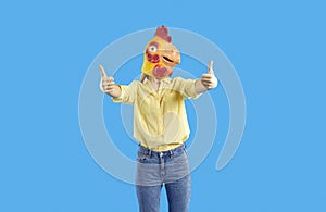 Cheerful funny and humorous woman in rubber mask of chicken shows thumbs up on blue background.