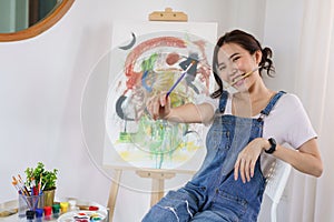 Creative of art concept, Young asian woman put paintbrush in mouth with artwork on canvas in studio