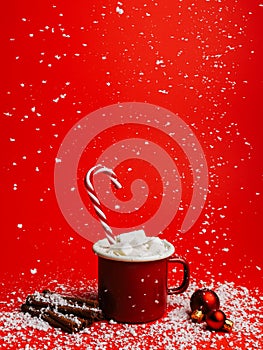 Creative Christmas composition with snow falling, tea cup, marshmallow drink and cinnamon sticks on red background. Minimal Xmas e
