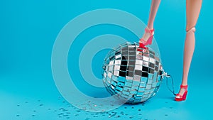 Creative Christmas party composition with doll leg in high heels and shiny disco ball on blue background. Minimal Xmas or New Year