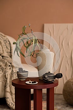 Creative composition of living room interior with wooden round coffee table, beige vase with leaves, tea in cup, pitcher, and