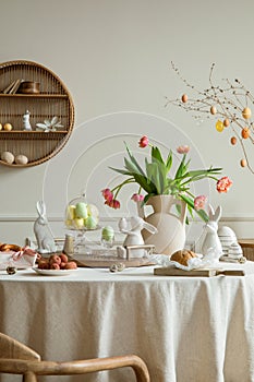 Creative composition of spring easter dining room interior with round table, vase with tulips, colorful eggs, wooden trace, bread