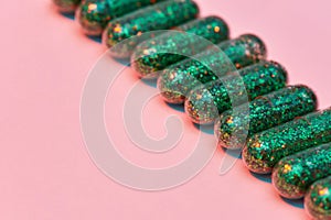 Creative concept with many green glitter pills lying diagonally in a row isolated on pastel pink background. Minimal
