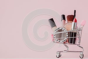 Creative concept with shopping trolley with makeup on a pink background. Perfume, sponge, brush, mascara, pencil, nail file, eye