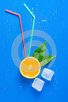 Ripe Juicy Cut in Half Orange Green Leaves Drinking Straws Melting Ice Cubes on Blue Background. Fresh Juices Summer Cocktails